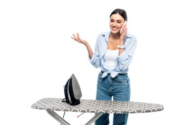 Ütü masasının yanında cep telefonuyla konuşan neşeli bir kadın beyaza izole edilmiş.