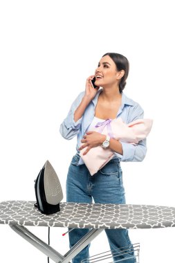 Bebek sahibi neşeli bir kadın akıllı telefonla konuşuyor. Ütü masasının yanında. Beyazlar içinde izole edilmiş.