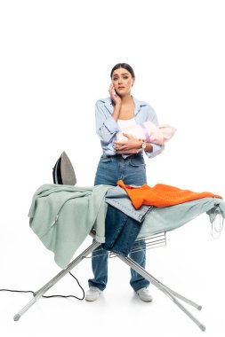 tired woman with infant baby talking on smartphone near ironing board on white background clipart