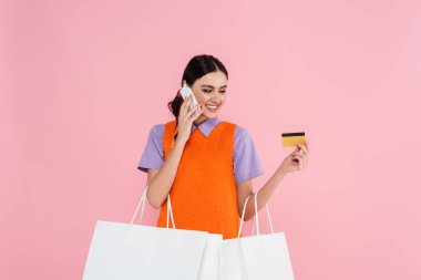 Kredi kartı ve alışveriş çantalarıyla mutlu bir kadın pembe bir telefondan konuşuyor.