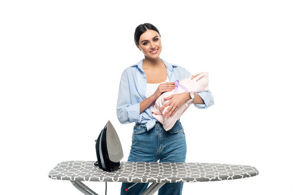 happy housewife smiling at camera while standing near ironing board with infant child isolated on white
