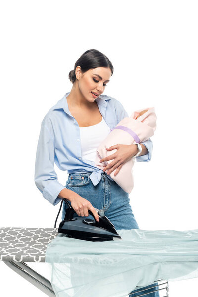 young mother holding newborn baby while ironing clothes isolated on white