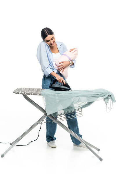cheerful woman talking on mobile phone while standing near ironing board with infant child on white background