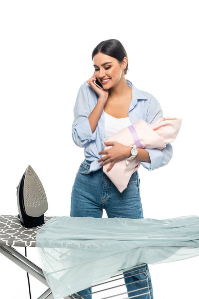 smiling woman holding newborn baby and talking on mobile phone near ironing board isolated on white