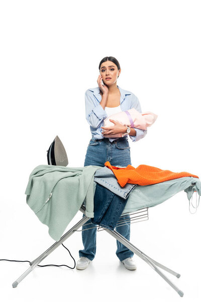 tired woman with infant baby talking on smartphone near ironing board on white background