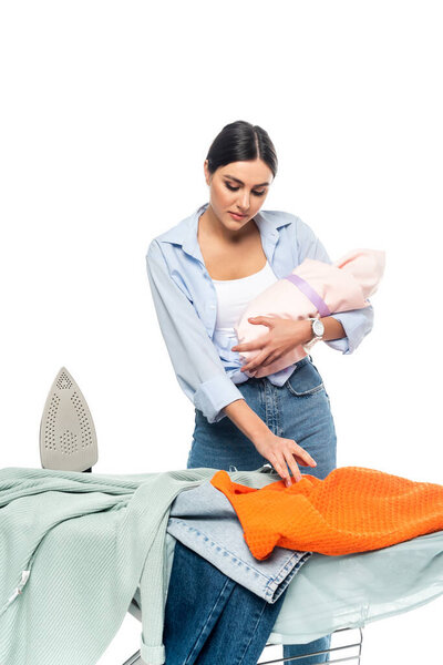 young mother holding newborn baby near clothes on ironing board isolated on white