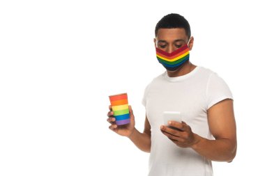 Koruyucu maskeli ve lgbt renginde kağıt bardaklı Afro-Amerikan bir adam beyaz telefondan izole edilmiş akıllı telefondan mesaj yolluyor.