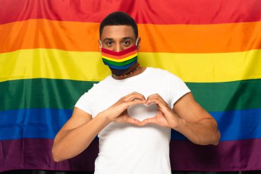 Afro-Amerikan adam gökkuşağı renginde tıbbi maske takıyor ve lgbt bayrağının arka planında kalp işareti gösteriyor.