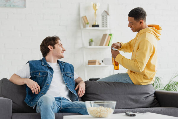 african american young man opening beer bottles and resting with friend in living room