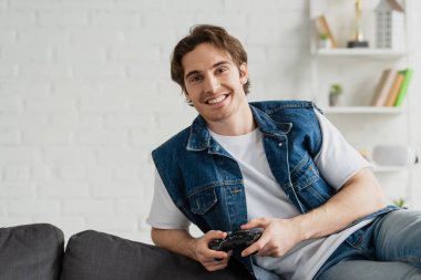 KYIV, UKRAINE - 22 Mart 2021: Genç gülümseyen adam rahat bir şekilde koltuğa oturmuş modern odada joystick ile poz veriyor