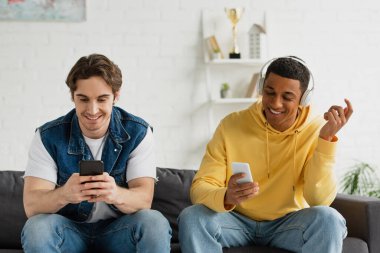 Akıllı telefonlu koltukta oturan ve oturma odasında kulaklıkla müzik dinleyen mutlu ırklar arası arkadaşlar.