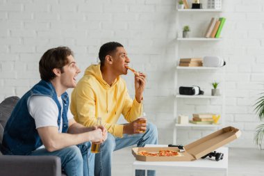 KYIV, UKRAINE - 22 Mart 2021: Afro-Amerikan genç adam oturma odasında arkadaşıyla pizza yiyor ve bira içiyor.
