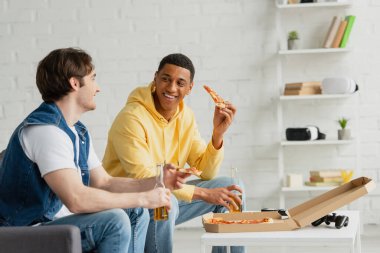 KYIV, UKRAINE - 22 Mart 2021: Afro-Amerikan gülümseyen adam oturma odasındaki kanepede arkadaşıyla pizza yiyor ve bira içiyor.