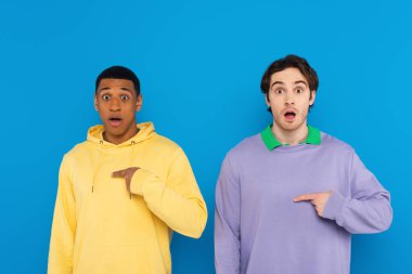 frustrated interracial friends looking at camera with open mouth and pointing to themselves with fingers isolated on blue clipart