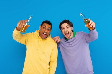 happy interracial friends lough and celebrating with bottles of beer isolated on blue clipart