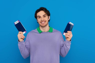 Elinde pasaportu olan mutlu genç bir adam mavi üzerine izole edilmiş uçak biletleri tutuyor.