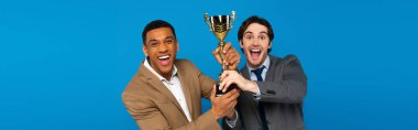 interracial friends in suits celebrating success with prize cup in hands isolated on blue, banner  clipart