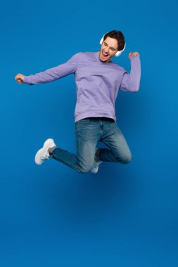 happy smiling man in violet sweatshirt enjoying music and jumping in headphones on blue background clipart