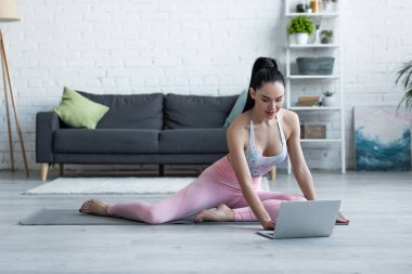 Spor kıyafetli esmer kadın yoga minderinde otururken dizüstü bilgisayara bakıyor.