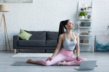 Evde laptopun yanında yoga minderi üzerinde meditasyon yapan genç bir kadın.