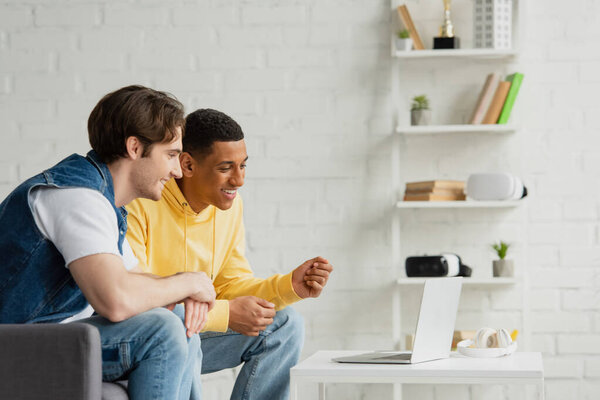 smiling interracial friends sitting together and watching at laptop in modern living room