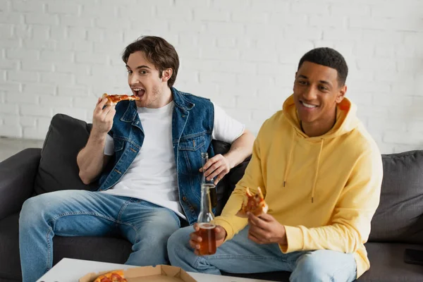 interracial friends enjoying pizza and beer while sitting on couch in modern loft