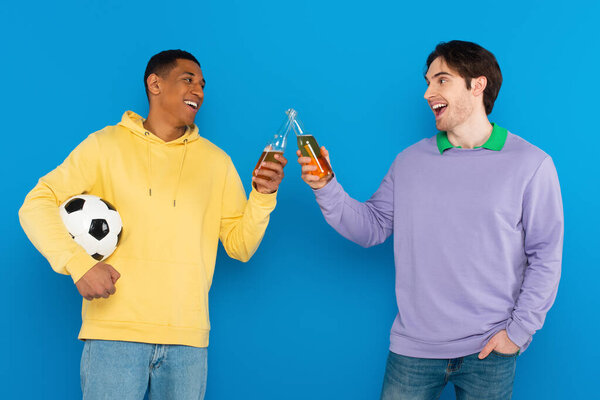 улыбающиеся межрасовые друзья, держащие футбол и тосты с бутылками пива, изолированными на голубом