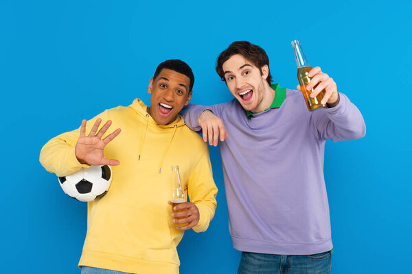 excited interracial sport fans holding football and bottle of beer isolated on blue