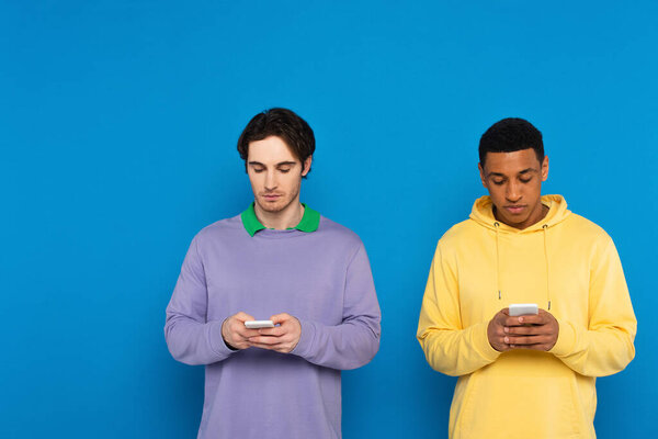 interracial friends staring at smartphones isolated on blue