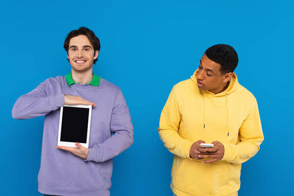 african american man looking at smiling friend with tablet isolated on blue