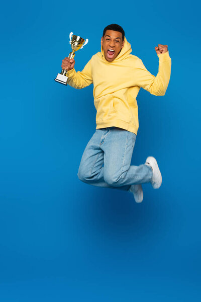 возбужденный африканский американец в желтой толстовке прыгает с футбольной чашкой на синем фоне