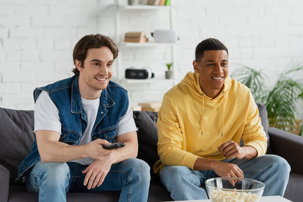 young interracial friends sitting on couch, clicking channels with tv remote controller and eating popcorn at home