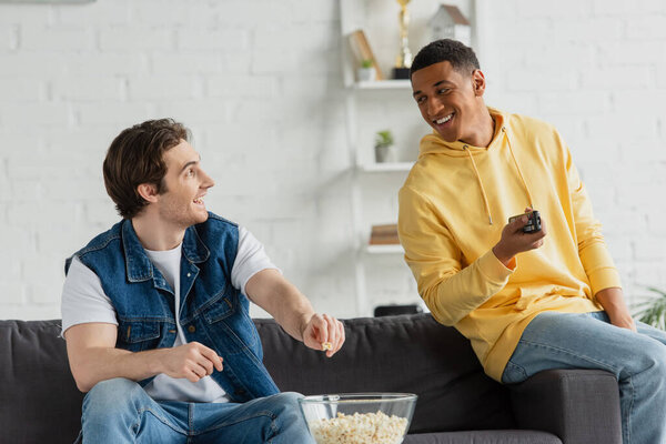 smiling african american man with friend clicking channels and eating popcorn in modern loft