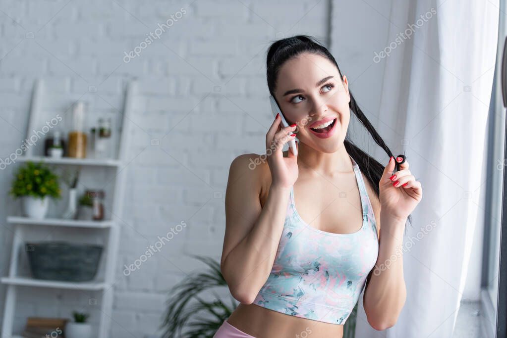 cheerful, coquettish woman in sports bra touching hair while talking on mobile phone at home