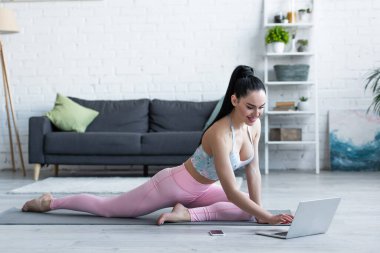 Evde yoga minderinde otururken dizüstü bilgisayara bakan gülümseyen kadın.