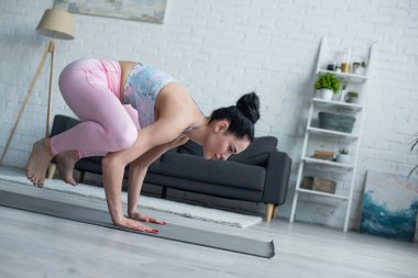Genç esmer kadın evde karga pozuyla yoga yapıyor.