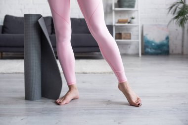 cropped view of barefoot woman in leggings standing near yoga mat at home clipart