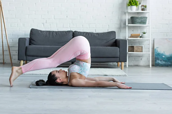 从侧面看穿着运动服的女性在家里瑜伽垫上练习犁地姿势 — 图库照片