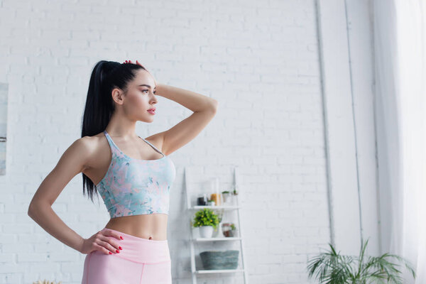 young sportswoman standing with hand on hip and looking away at home