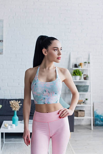 sportive woman in sports bra standing with hand on hip and looking away at home