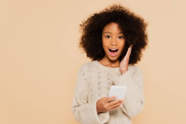 Heyecanlı Afrika kökenli Amerikalı genç kız. Yüzüne yakın, bejde izole edilmiş akıllı telefonu tutuyor.