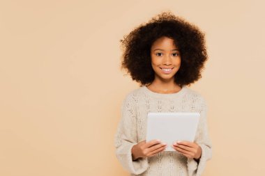 Afro-Amerikan reşit olmayan kız bej renkli ellerde tabletle kameraya bakıyor.