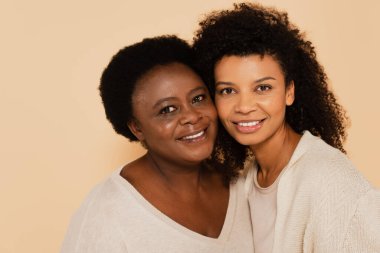 Gülümseyen Afrikalı Amerikalı yetişkin kız ve orta yaşlı anne bej renginde yanak yanağa sarılıyorlar.