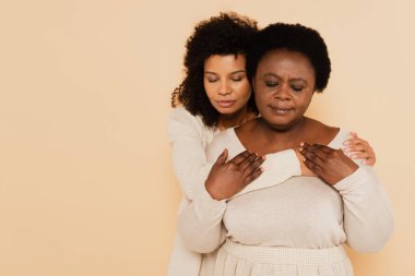 Afrikalı Amerikalı orta yaşlı anne ve yetişkin kız bej rengi gözlerle sarılıp el ele tutuşuyorlar.