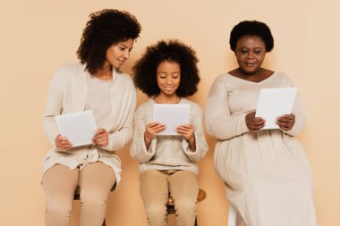 Afro-Amerikan kızı, torunu ve büyükannesi bej arka planda oturup tabletlere bakıyor.