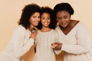 african american daughter, granddaughter and grandmother hugging and looking at camera on beige background clipart