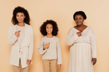 african american daughter, granddaughter and grandmother pointing with fingers to themselves on beige background clipart