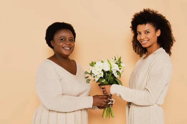 smiling african american adult daughter and middle aged mother with bouquets of daisies looking at camera on beige background