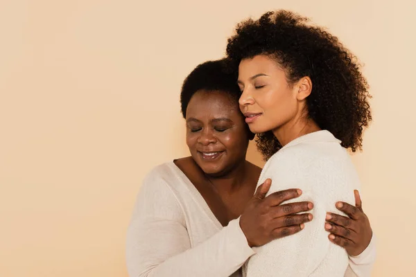 portrait of african american adult daughter and middle aged mother hugging with closed eyes isolated on beige