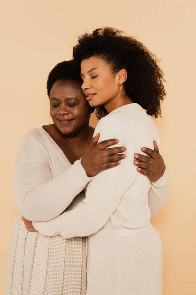 african american adult daughter and middle aged mother hugging with closed eyes isolated on beige
